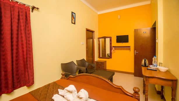 A hotel room with a bed, a wooden nightstand, a chair, a mirror, and a red curtain. - Malar Residencey (Puducherry)