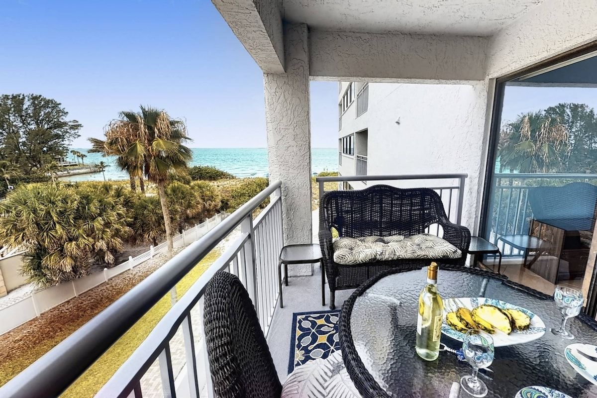 A balcony with a view of the sea, wicker furniture, a table with a bottle and glasses, and a patterned rug.