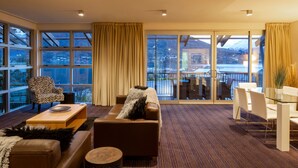 Habitación de hotel moderna con un amplio ventanal corredera que da a un balcón con vistas a una montaña nevada y una masa de agua. En el interior, hay un sofá de cuero marrón, un sillón con estampado de cebra y una mesa de comedor de vidrio con sillas blancas.