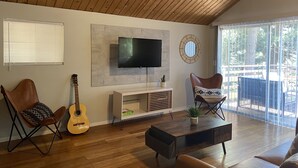 Un salon avec un plafond en bois, un téléviseur à écran plat fixé au mur, une guitare, deux fauteuils, une table basse et une grande fenêtre avec des stores.