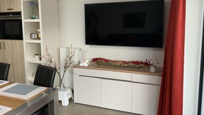 Ett modernt vardagsrum med en platt-TV som är monterad på väggen, ett matbord med bordsunderlägg, ett vitt skåp och en liten pall.