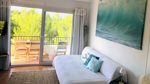Una habitación con cama, un cuadro de una ola, un balcón con sillas y vista a árboles.