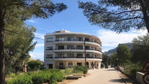 Un edificio de varios pisos con balcones, una calle pavimentada en primer plano y montañas al fondo.