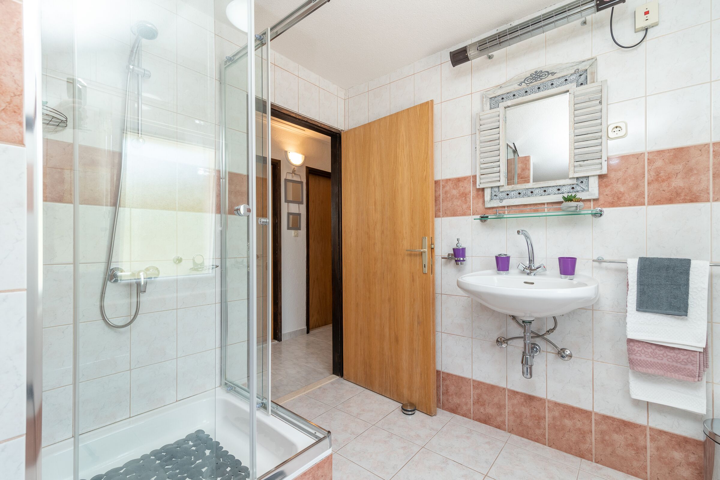 A bathroom with a glass shower enclosure, a sink with a mirror above it, and a wooden door leading to another room.