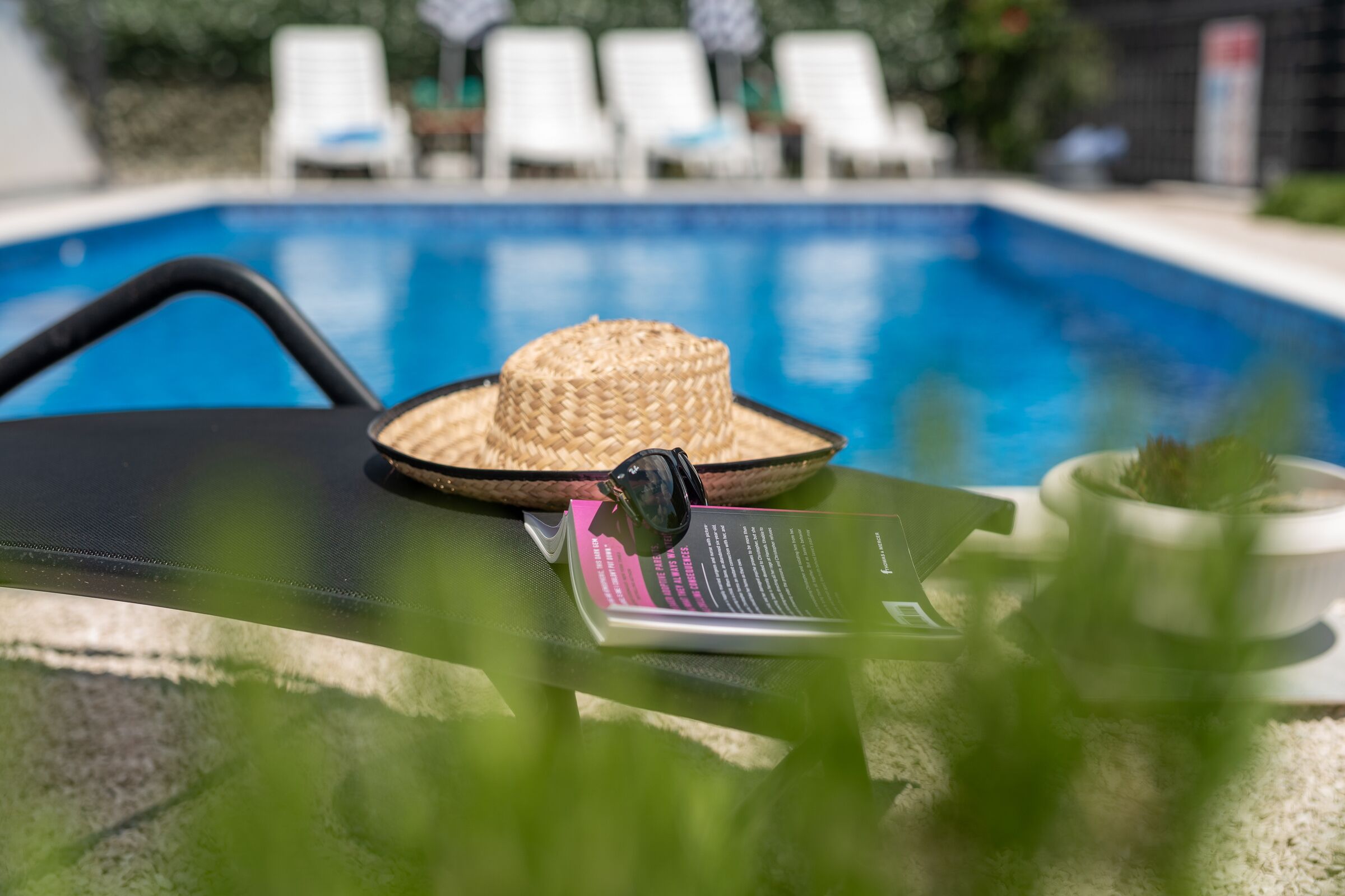 A poolside area with a lounge chair, a straw hat, sunglasses, a book, and a potted plant.