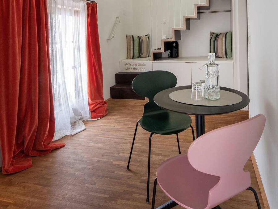 A room with a wooden floor, a round table with chairs, a staircase, and a kitchen area in the background.