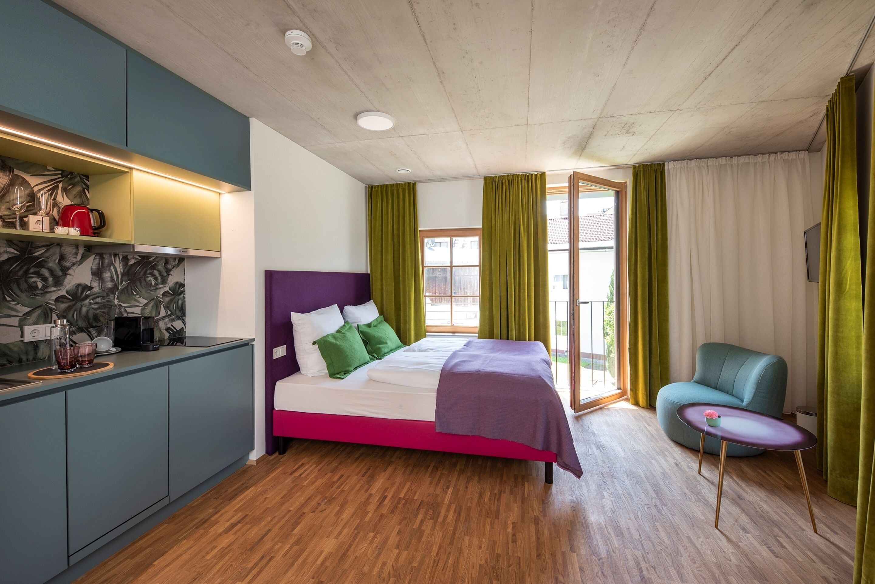 A modern bedroom with a bed, a chair, a small table, and a kitchenette.