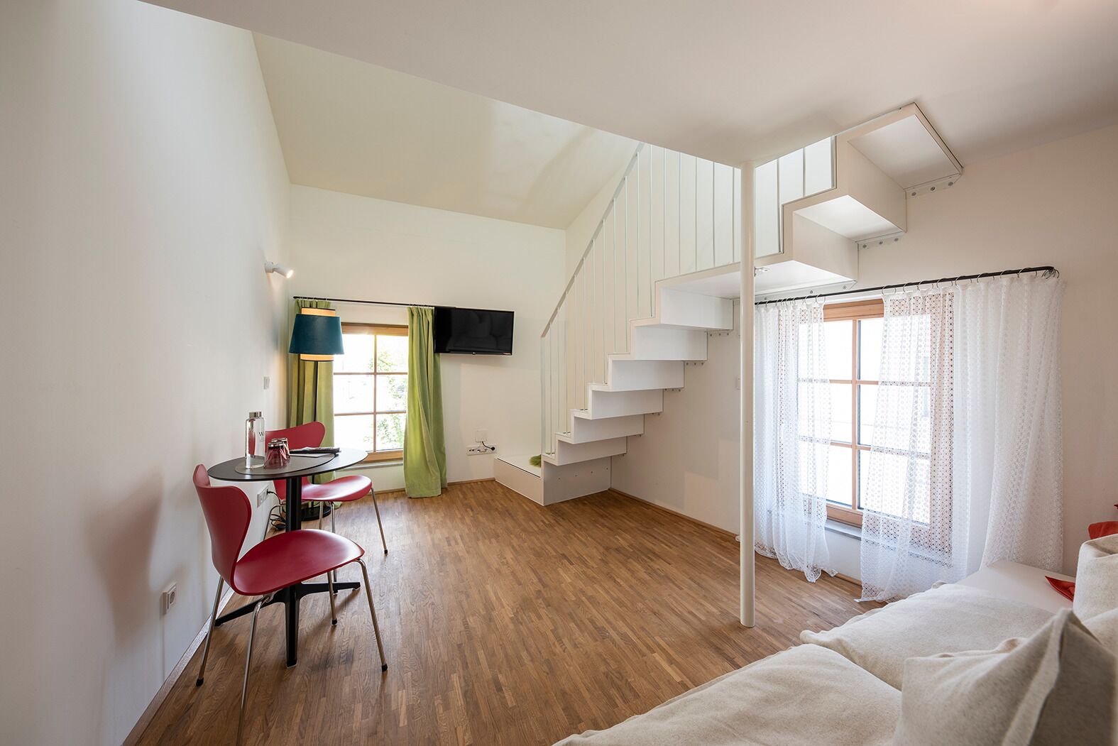 Una habitació moderna amb terra de fusta, una zona de menjador amb taula i cadires, una escala i un llit.