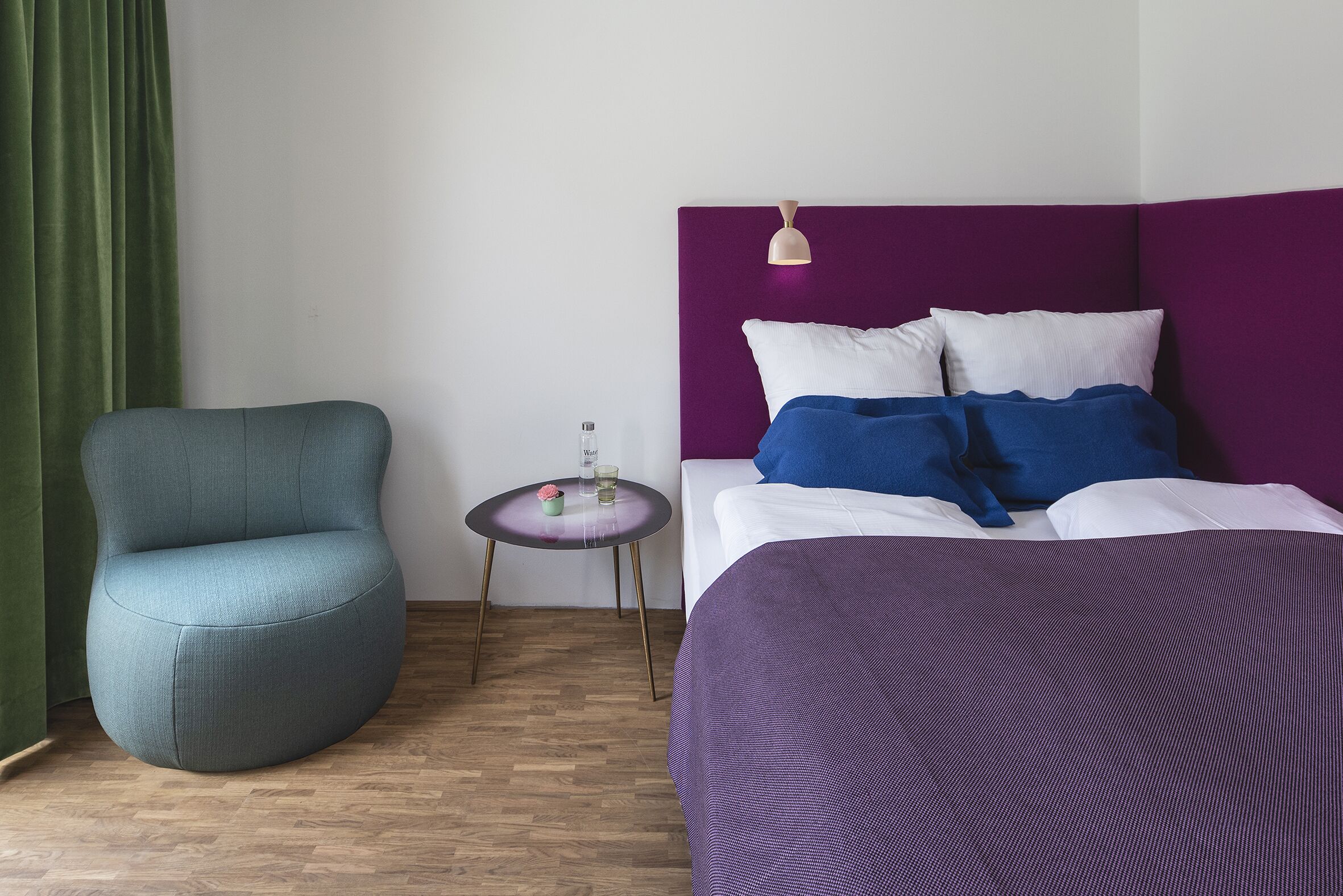 Een kamer met een bed, een stoel, een klein tafeltje en een groen gordijn.