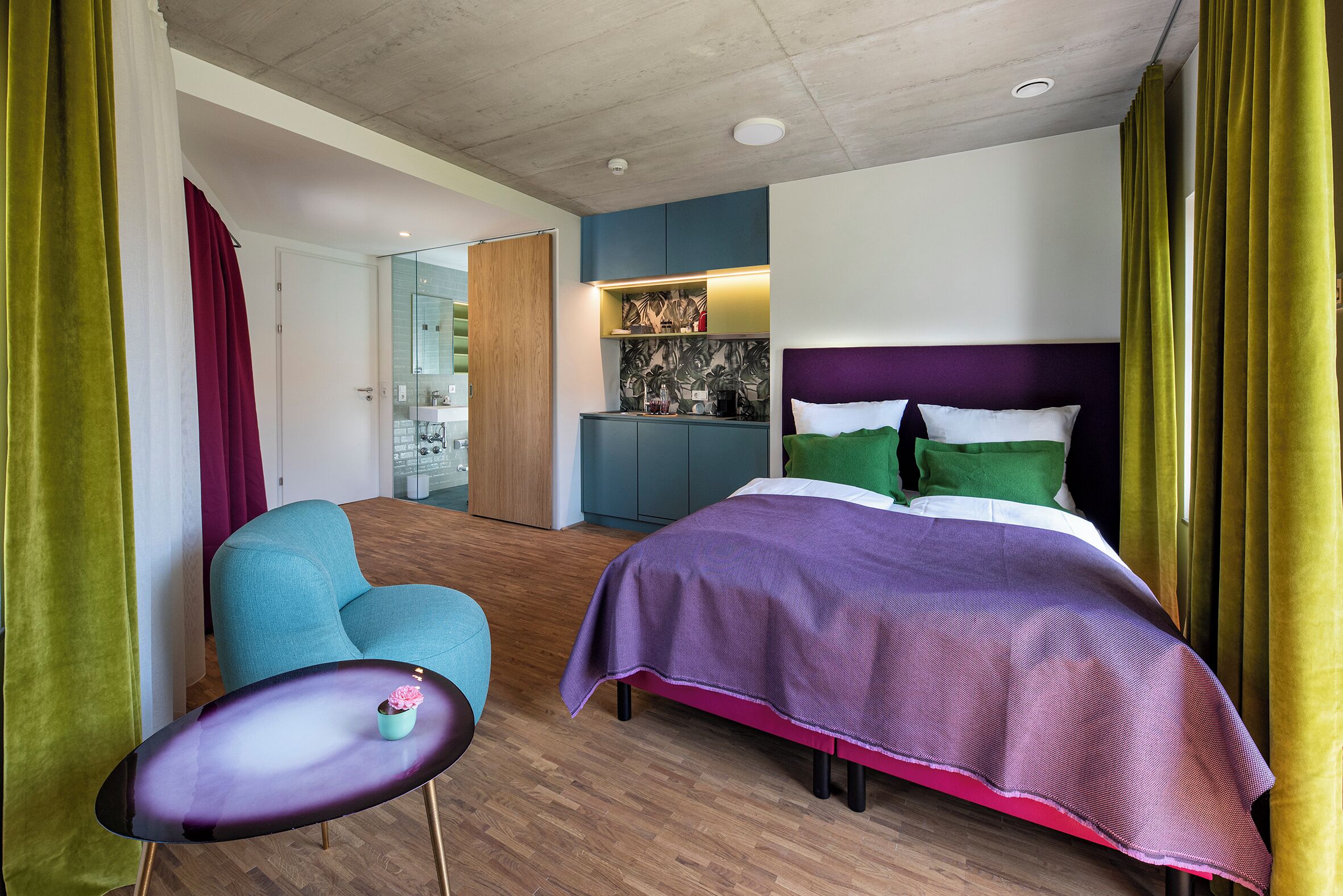Quarto de hotel moderno com cama, área de estar, cozinha compacta e banheiro.