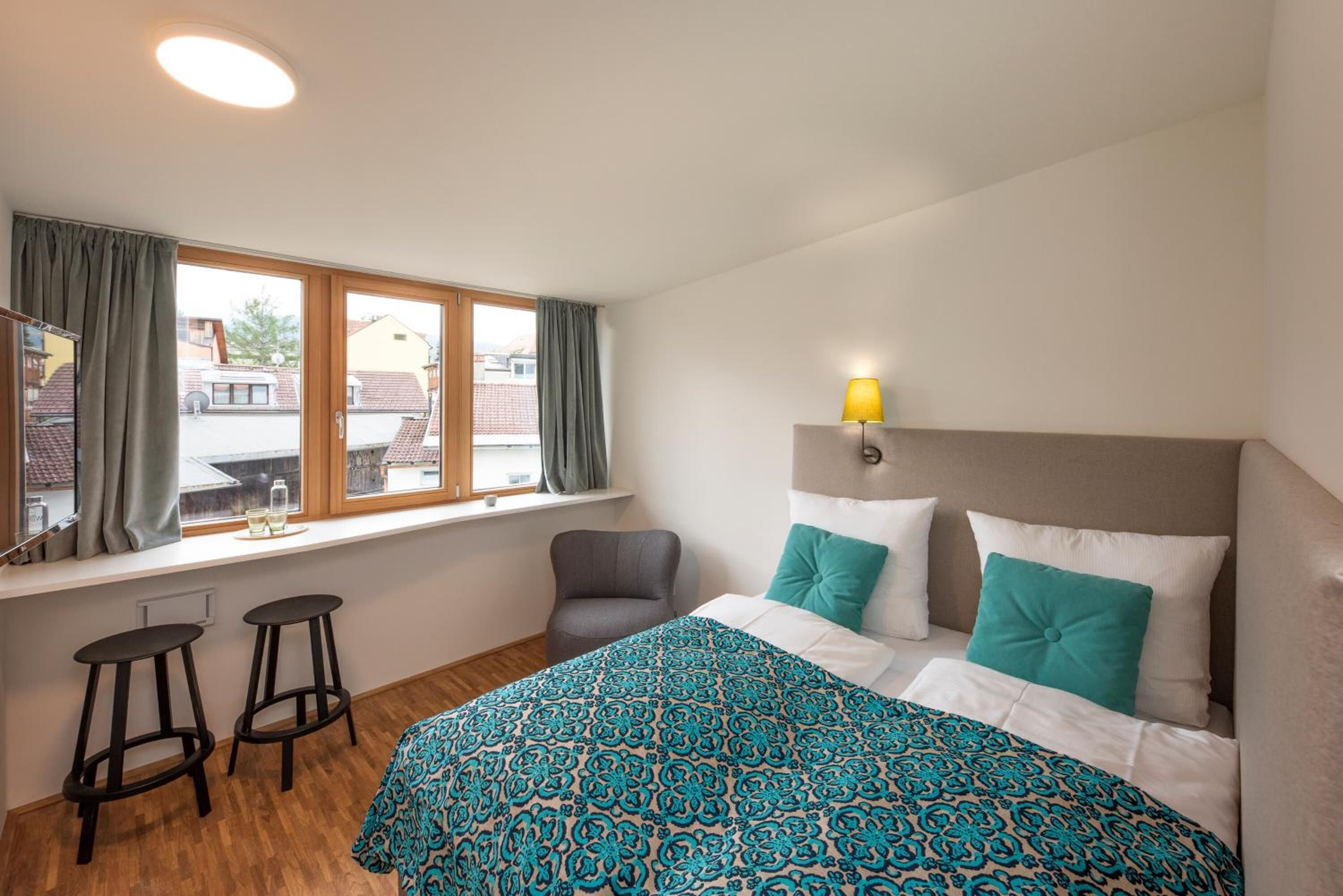 A hotel room with a bed, a window with curtains, a chair, and two stools.