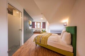 Quarto moderno com cama verde, piso de madeira, área de estar com uma cadeira e uma mesa, e vista para a cidade através de grandes janelas.