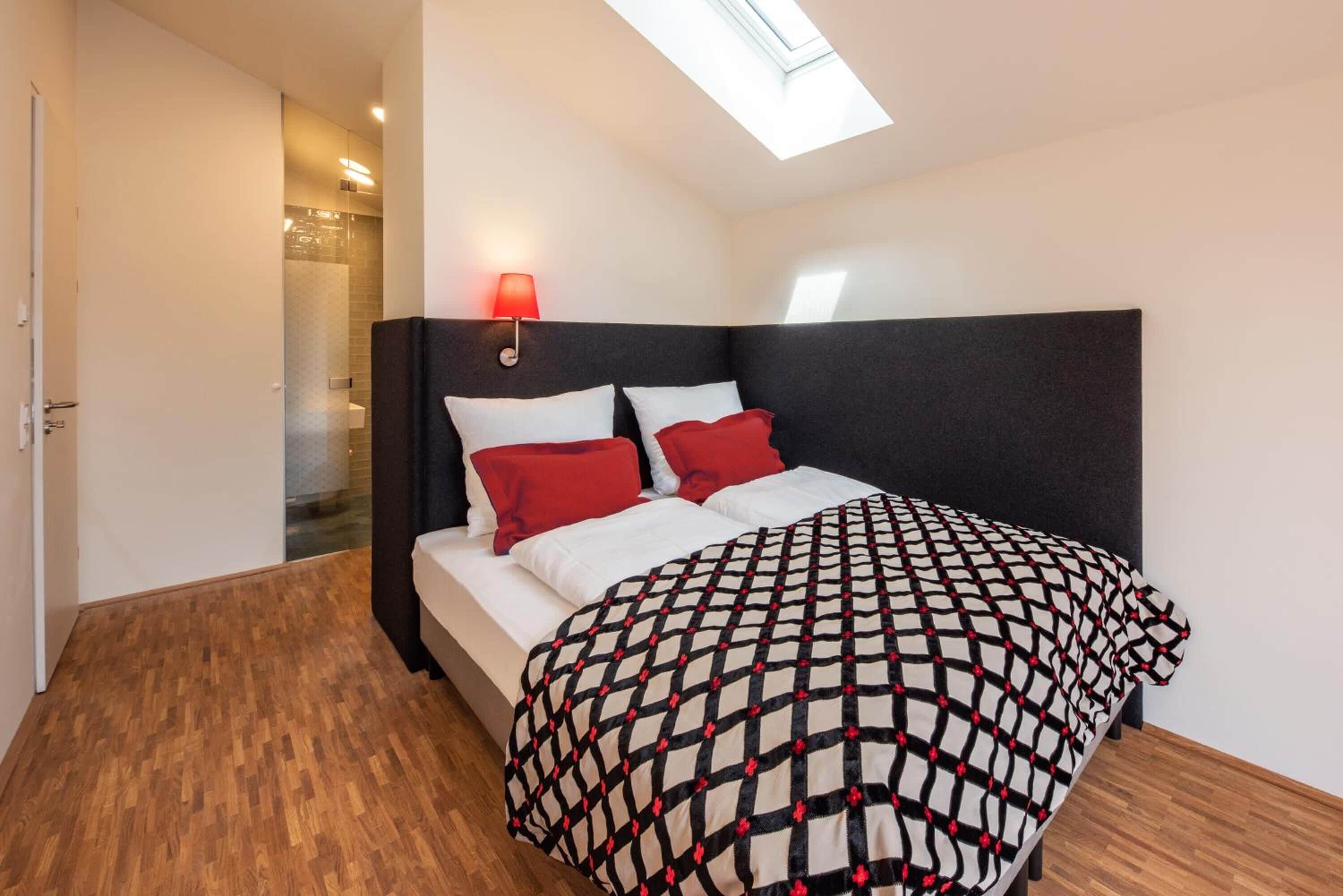 Une chambre avec un puits de lumière, un lit recouvert d’une couvre-lit à motifs et un lampadaire rouge fixé au mur.