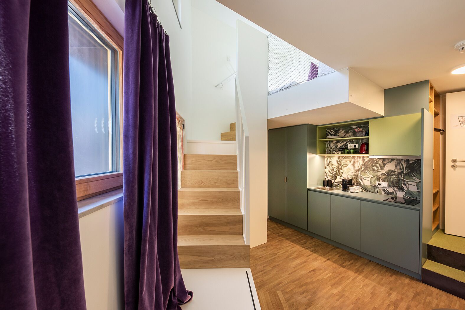 A modern kitchen with green cabinets, a wooden staircase, and a window with purple curtains.