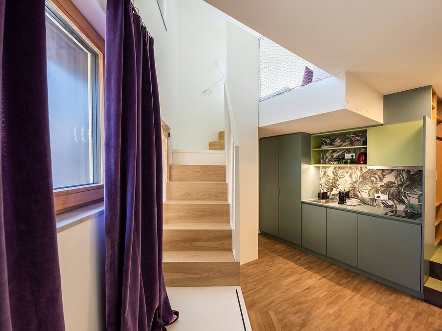 A modern kitchen with green cabinets, a wooden staircase, and a window with purple curtains.