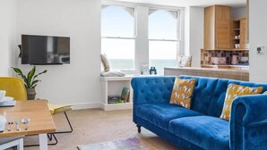 A modern living room with a blue sofa, a wooden dining table, and a TV mounted on the wall. - Stylish Flat with Stunning Sea Views, Ilfracombe (Ilfracombe)