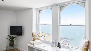 A room with a large window overlooking the sea, a flat-screen TV mounted on the wall, a small table with a plant, and a bench with pillows and a sign. - Stylish Flat with Stunning Sea Views, Ilfracombe (Ilfracombe)