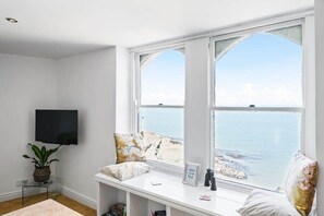 Habitación con un ventanal que da al mar, un televisor de pantalla plana en la pared, una mesita con una planta y un banco con almohadones y un letrero.