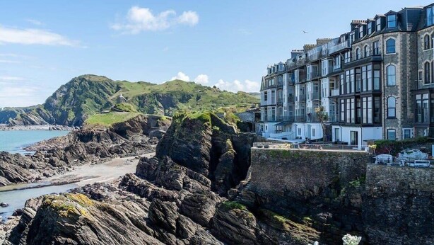A coastal building with a stone facade and multiple windows, situated on a rocky shoreline with a view of the sea and a hill in the background. - Stylish Flat with Stunning Sea Views, Ilfracombe (Ilfracombe)
