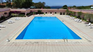 A rectangular pool with a tiled deck, surrounded by white lounge chairs and a view of the sea. - Blue Bay Apartment Baja Sardinia (Arzachena)