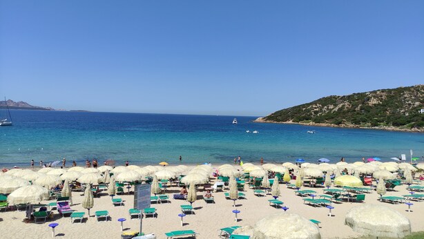 A beach with rows of sunbeds and umbrellas, a clear blue sea, and boats in the distance. - Blue Bay Apartment Baja Sardinia (Arzachena)