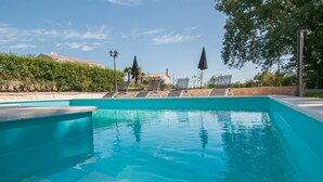 Une piscine d’un bleu éclatant, dotée de chaises longues et de parasols, entourée de verdure et avec des bâtiments en arrière-plan.