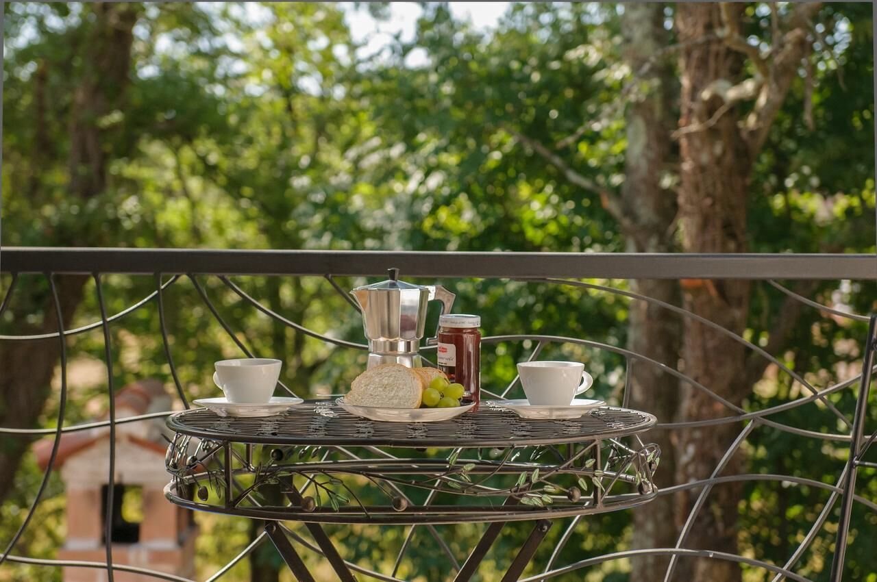 Una mesa metálica con una cafetera, dos tazas, un tarro de mermelada y un plato con pan y uvas en un balcón con un árbol al fondo.