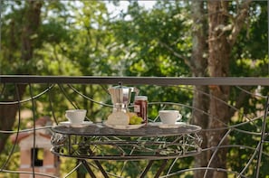 A metal table with a coffee pot, two cups, a jar of jam, and a plate with bread and grapes on a balcony with a tree in the background.
