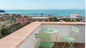 A balcony with two green chairs and a small table, overlooking a coastal cityscape with buildings and a clear view of the sea. - Terramare Guesthouse (Procida)