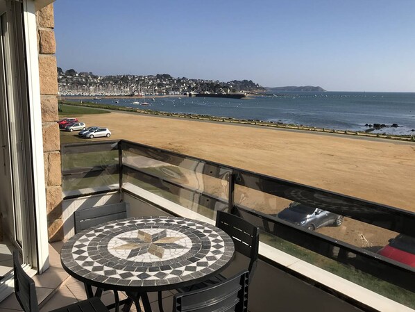 Een balkon met een betegelde tafel en stoelen, met uitzicht op een strand en een jachthaven.