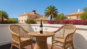 Un balcon avec des fauteuils en osier et une bouteille de vin, donnant sur un bâtiment avec un toit en tuiles et une végétation luxuriante.