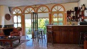 A spacious living area with a wooden bar and stools, a fireplace, and large windows with decorative arches.