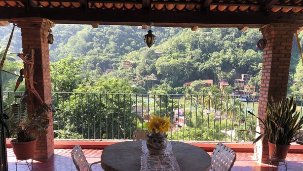 Ein Balkon mit rundem Tisch und Stühlen, Blumentöpfen und Blick auf eine üppige grüne Landschaft mit Gebäuden in der Ferne.