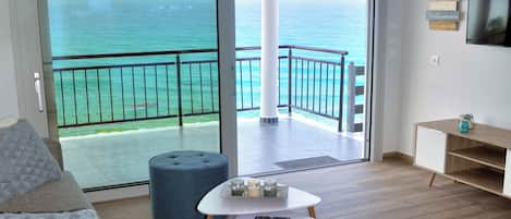 A spacious living area with a balcony overlooking the sea, a modern sofa, a small table with candles, and a TV mounted on the wall.