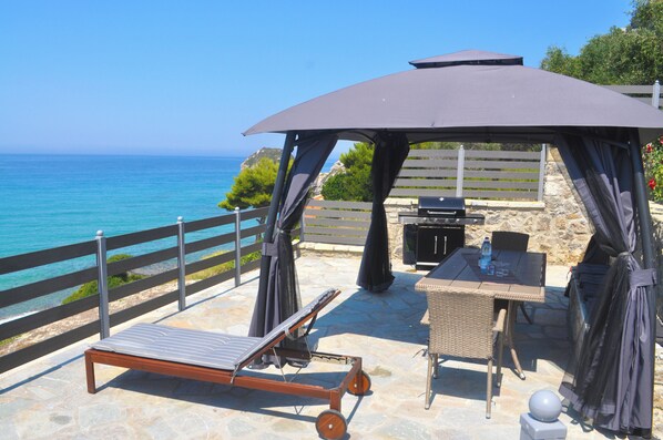 A patio with a sun lounger, a table, and a grill, overlooking the sea.