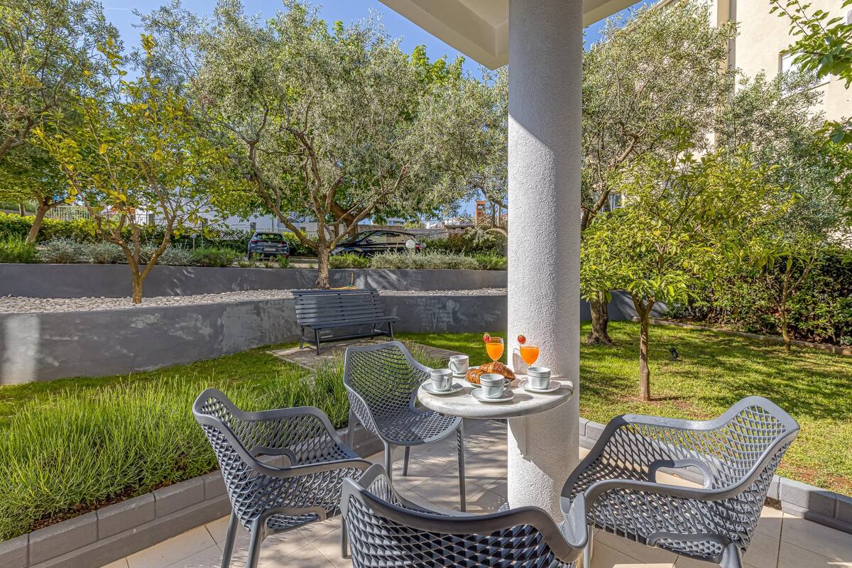 Eine Terrasse im Freien mit einem für zwei Personen gedecktem Tisch, umgeben von Stühlen und Blick auf einen Garten mit Bäumen und einer Bank.