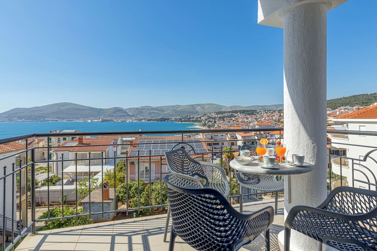 Un balcón con sillas y mesa de metal, con vistas a un pueblo costero con edificios y un mar azul cristalino.