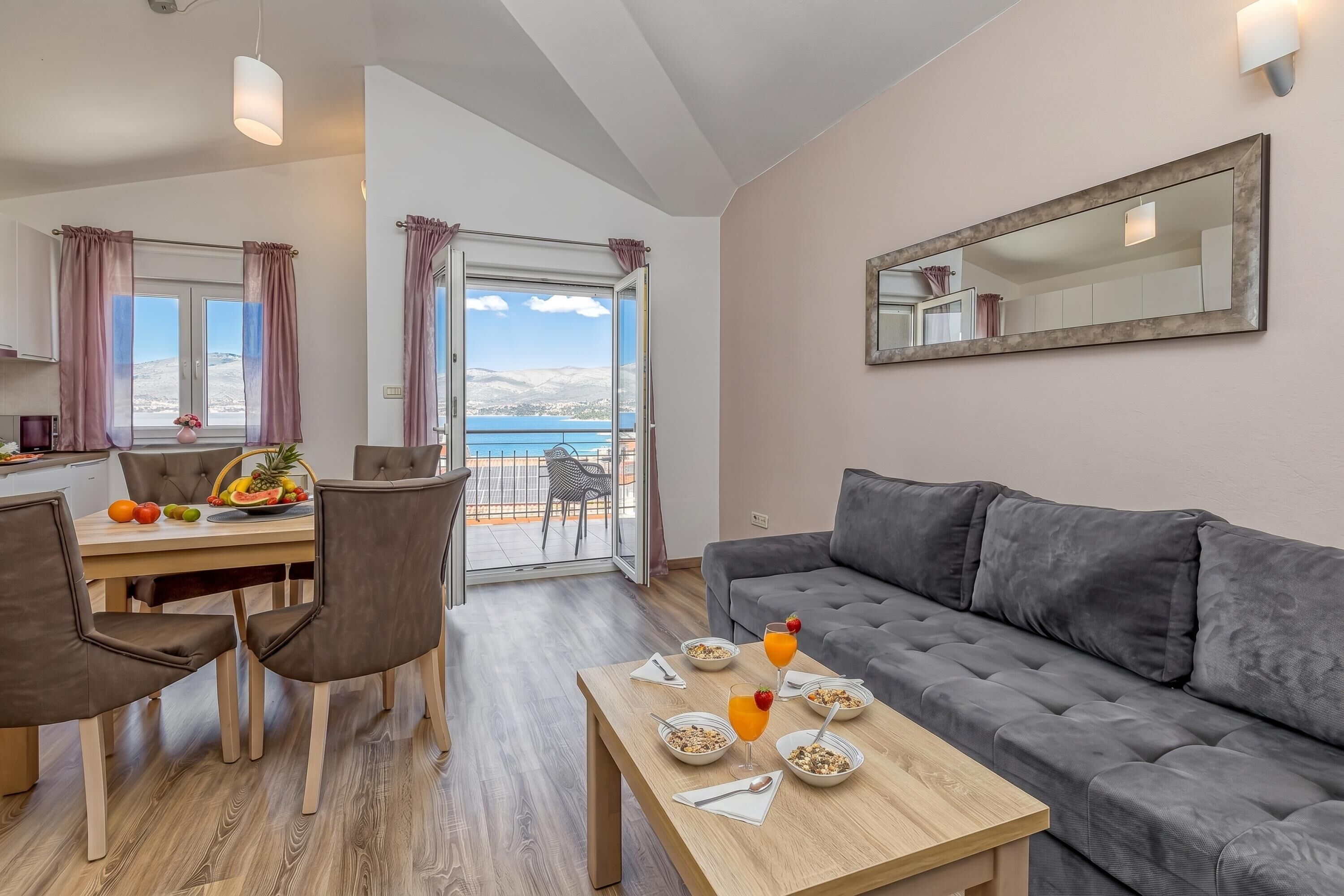 Ein modernes Wohnzimmer mit grauem Sofa, einem Esstisch mit Stühlen, einer Frühstücksecke und einem Balkon mit Blick.
