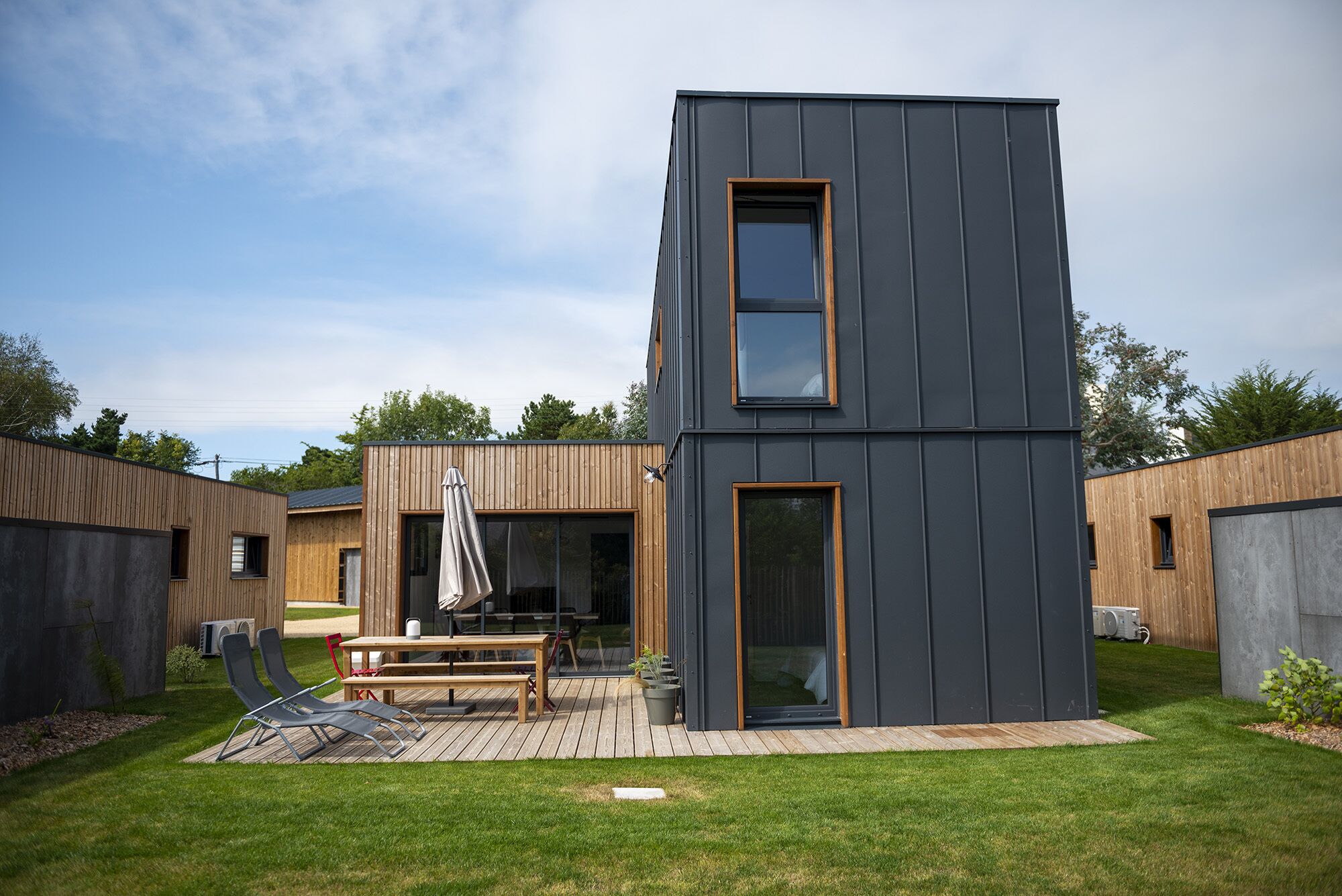 Een modern gebouw met twee verdiepingen, een plat dak, grote ramen en een houten terras met zitplaatsen.