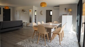 Een moderne woonkamer met een bank, eettafel en stoelen. Er is een groot raam en een kast met een plant erboven.