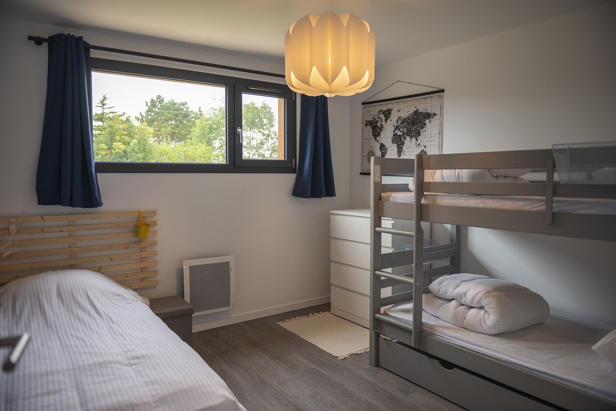 A bedroom with a bunk bed, a single bed, a window with curtains, a hanging light fixture, and a world map on the wall.