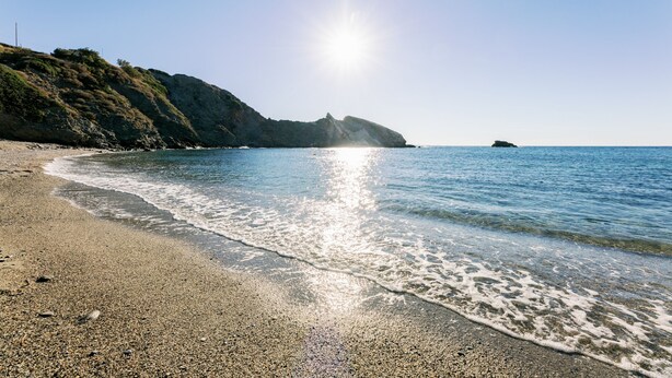 Ein Strand mit Kiesstrand, klarem Wasser und einer in der Ferne liegenden Felsformation im hellen Sonnenschein.