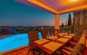 Un espace repas aménagé au bord de la piscine, offrant une vue sur la mer et la ville au crépuscule.