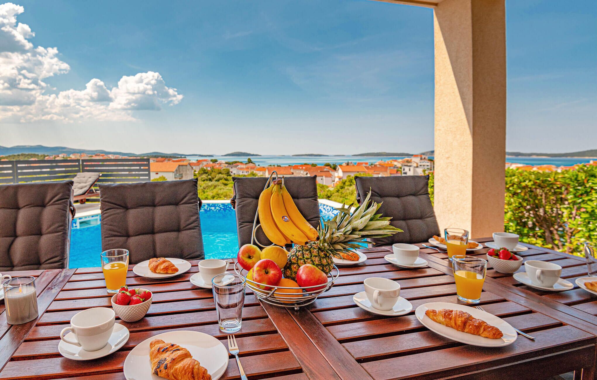 A breakfast table with croissants, fruits, and juice, overlooking a pool and a scenic view.