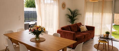A dining area with a wooden table and white chairs, a red sofa, and a small side table with a candle and a plant.