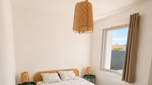 Une chambre à coucher avec un lit, deux tables de chevet, une fenêtre avec des rideaux et un luminaire suspendu.