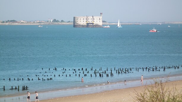 Un paysage côtier avec un bâtiment en pierre d’époque, des bateaux et une plage de sable.