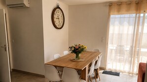 Un coin repas avec une table en bois, des chaises blanches, une horloge et une fenêtre ornée de rideaux légers.