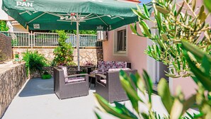 Ein Außenbereich mit einer Terrasse, Korbmöbeln, einem grünen Sonnenschirm und Blumentöpfen.