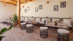 Un patio soleado con techo de madera, un conjunto de sofás, mesas redondas y plantas en macetas.